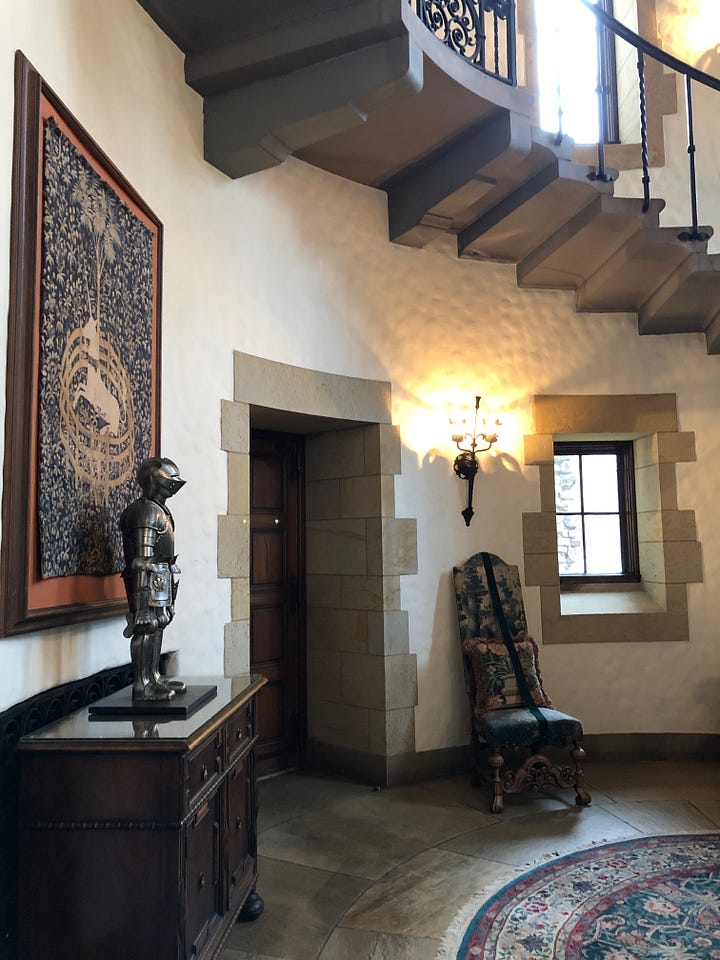 Four views of a circular stair well. From top-left to bottom-right: view of a circular carpet and two-seater sofa below a wrought-iron staircase with windows; a sidetable on which a suit of armour rests, with a carpet showing a unicorn on the wall and a tall-backed chair near a window; a view up the staircase showing wrought-iron light fittings; windows, a curving ironwork staircase, and hanging ironwork light fittings.