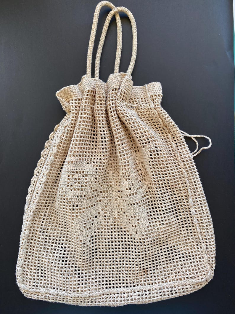 May include: A beige crochet drawstring bag featuring a mesh grid pattern throughout the body and a solid crochet butterfly motif centered on the front. The bag includes two long, thin cord handles and a drawstring closure at the top opening. May include: A beige crochet drawstring bag featuring a mesh grid pattern throughout the body and a solid crochet butterfly motif centered on the front. The bag includes two long, thin cord handles and a drawstring closure at the top opening.