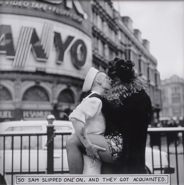 A sailor and a person in a feathered boa share a kiss in an urban setting under a large "Sanyo" sign on the building behind them. The mood is intimate and bold. There is a car behind the fence behind them. The sailor is kissing the drag queen from the previous frames. The caption on the bottom center of the image says 'So Sam slipped one on, and they got acquainted.' The photo is in black and white. The sailor is holding the drag queen's leg, which is mostly bare. She is wearing a dark coat with some black gloves. The sailor is wearing a white shirt, white pants, with a white hat.