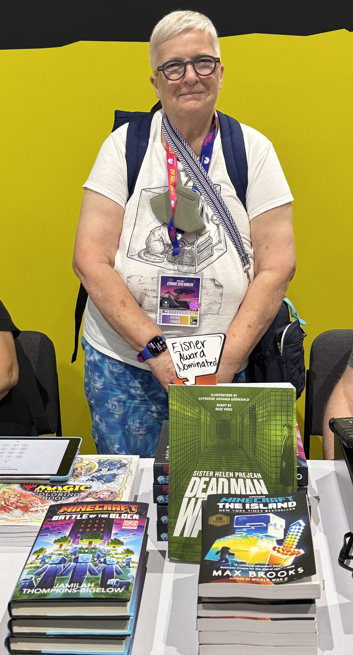 Rose Vines stands proudly behind Dead Man Walking: Graphic Edition at the Random House booth during San Diego Comic-Con. A handwritten "Eisner Award Nominated" sign rises above the book, which is displayed prominently among stacks of graphic novels. Wearing her Comic-Con professional badge, she smiles for the camera against the booth's bright yellow backdrop.