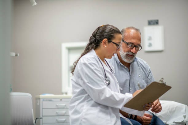 Medical Staff Prepare Their Infectious Control Attire stock photo A female doctor sits next to an elderly male patient during the Covid-19 outbreak.  They are reviewing the patient chart together. clinical trials patients stock pictures, royalty-free photos & images