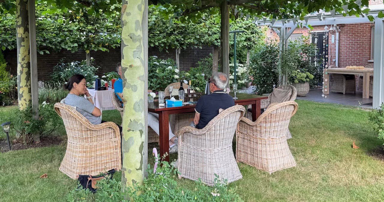Several people sit around a wooden table under a leafy pergola in a lush garden, engaged in conversation. They are seated in wicker chairs, with drinks and snacks on the table. In the background, more wicker chairs and a table are visible near a brick house, surrounded by neatly kept greenery and flowering plants.
