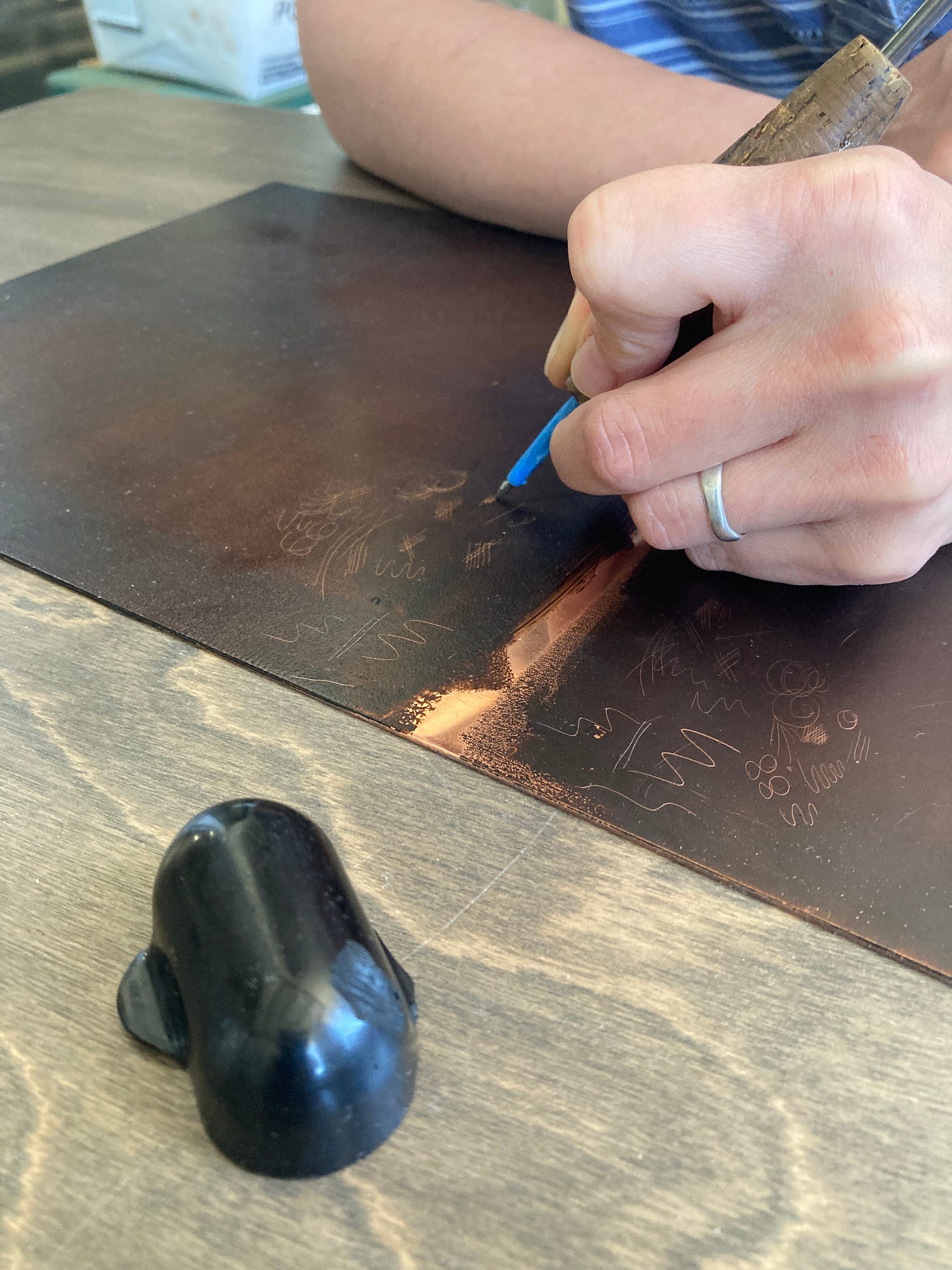 A hand holding a scribe draws lines and scribbles on a matte brown surface. Where the scribe has drawn, there is shiny copper. A ball of hard ground rests in the foreground.