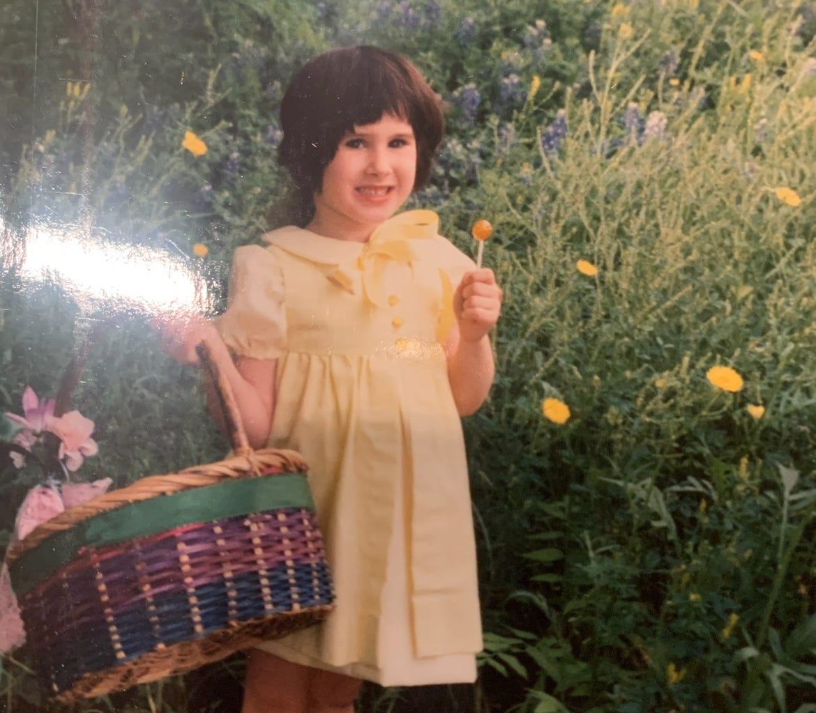 Lyric, as a very small child, in a field holding an easter basket. Lyric is wearing a yellow easter dress and is smiling but doesn’t look happy.
