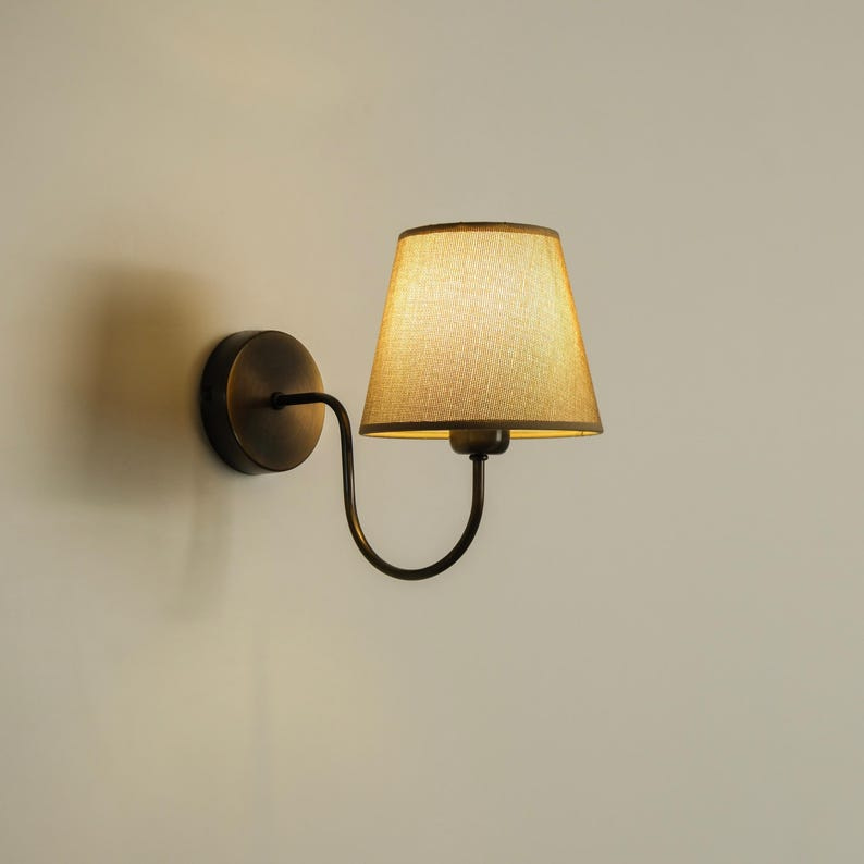 May include: A wall-mounted sconce with a bronze finish. The lamp features a curved arm supporting a beige lampshade. The shade has a tapered shape, and the fixture is mounted on a circular base. The design is simple and elegant. May include: A wall-mounted sconce with a bronze finish. The lamp features a curved arm supporting a beige lampshade. The shade has a tapered shape, and the fixture is mounted on a circular base. The design is simple and elegant.