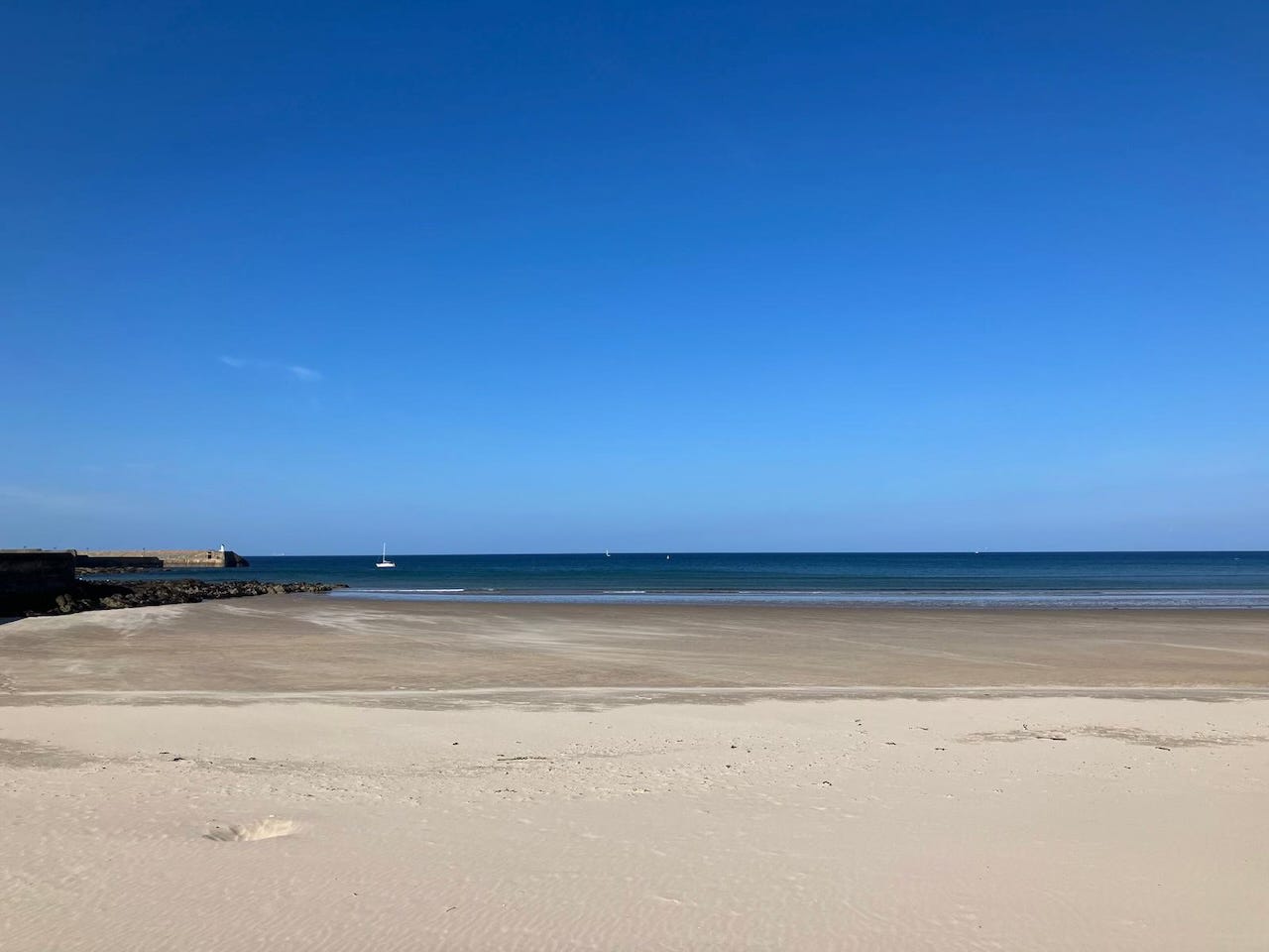 A wide sandy beach on the north coast of Scotland under a clear blue sky, with calm waves meeting the shore and a single white sailboat visible on the horizon. The scene feels open, peaceful, and quiet, inviting reflection and creativity.