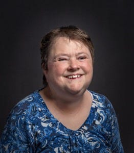 Abbie wears a blue and white V-neck top with different shades of blue from sky to navy that swirl together with the white. She has short, brown hair and rosy cheeks and smiles at the camera against a black background.