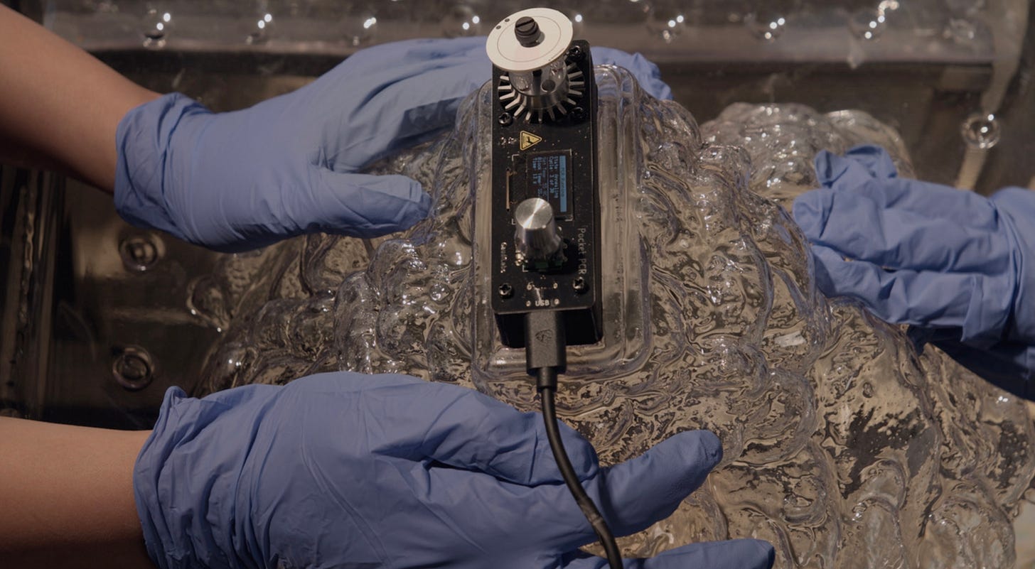 Three latex gloved hands are touching a clear blob connecting to a device implanted on the surface.