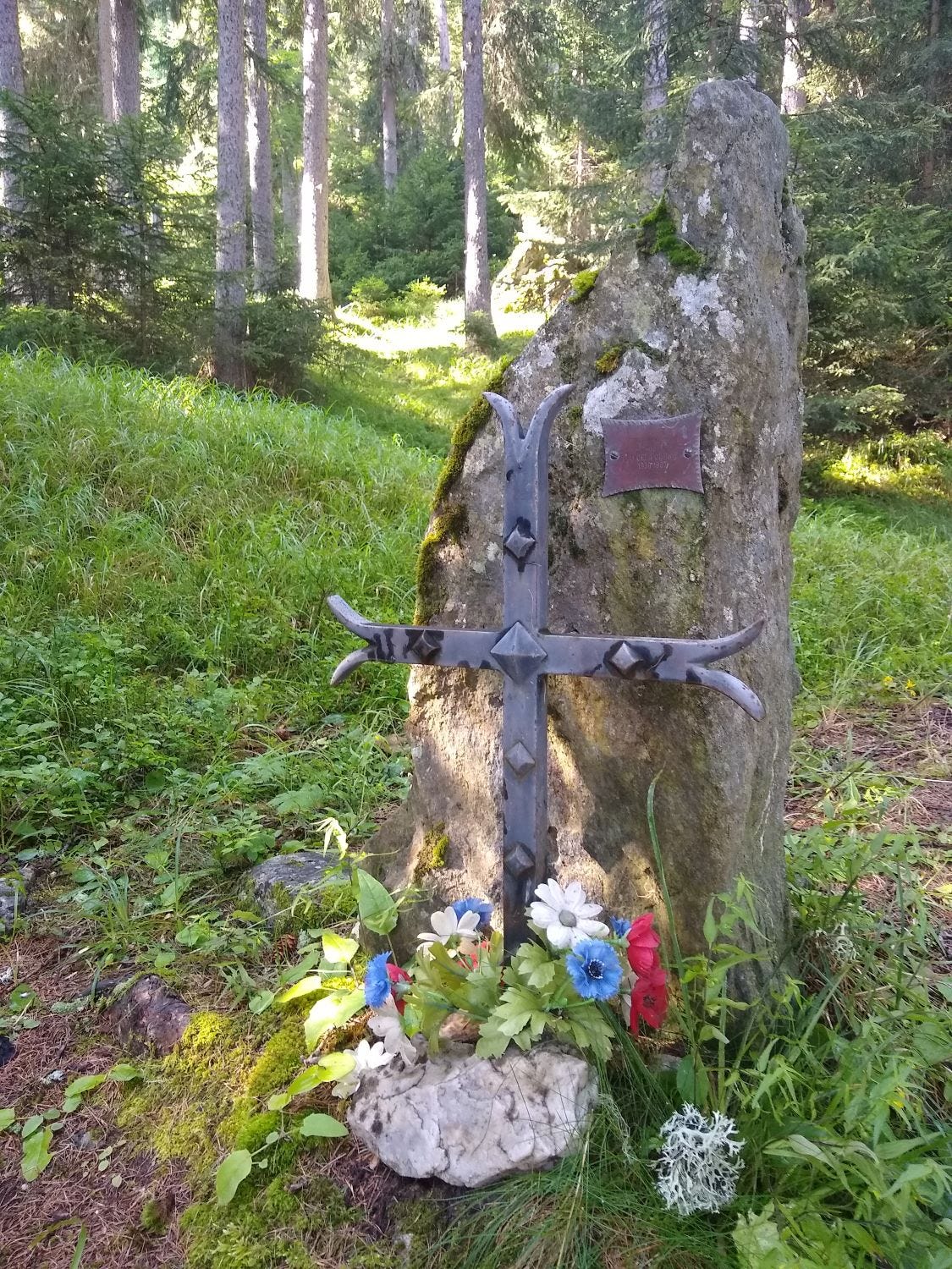 A gravestone, marked with flowers, in a peaceful woodland setting.