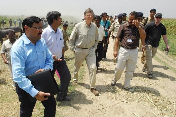 Bill Gates strides on a dirt path in a field in India. Many people surround him. Bill Gates strides on a dirt path in a field in India. Many people surround him.