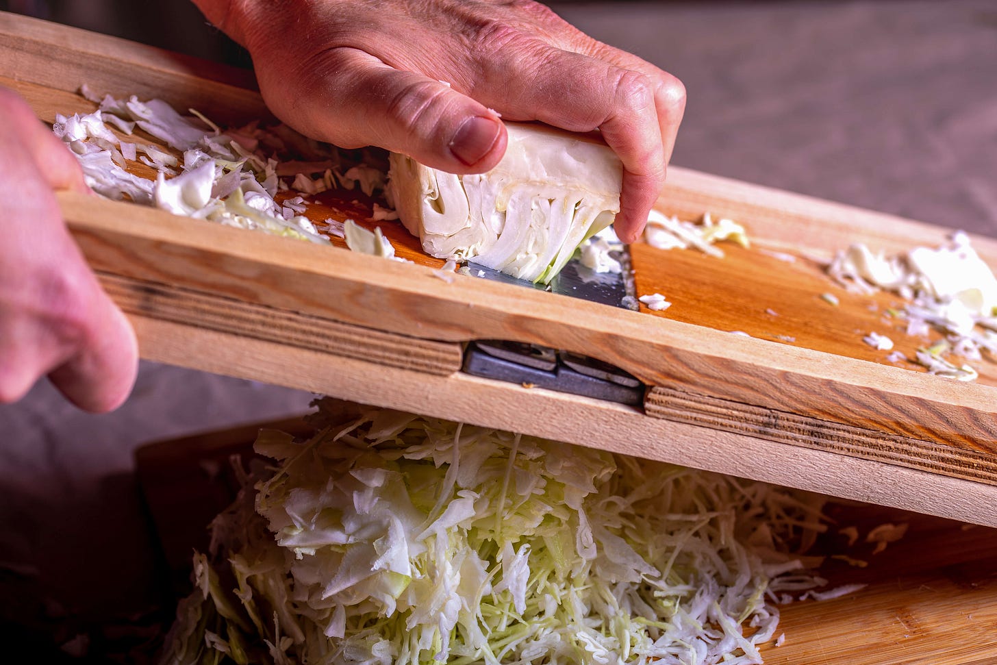 Hands slicing fresh white cabbage on a wooden mandoline, fine shreds falling into a bowl below.