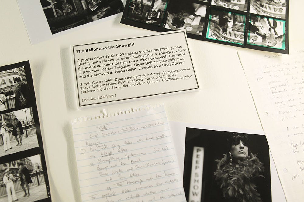 Black and white photo featuring photos, handwritten notes, and a typed card labeled "The Sailor and the Showgirl." The scene conveys creativity. It shows photographs and notes from Tessa during her time working on The Sailor and the Showgirl. The rest of the typed card reads: 'A project dated 1992-1993 relating to cross dressing, gender identity and safe sex. A 'sailor' propositions a 'showgirl, where the use of condoms for safe sex is also advocated. The sailor is a woman, Nerina Ferguson, Tessa Boffin's then girlfriend, and the showgirl is Tessa Boffin, dressed as a Drag Queen Smyth, Cherry 1996: Dyke! Fag! Centurion! Whore! An appreciation of Tessa Boffin' in Home, Peter and Lewis, Reina (ed) Outlooks: Lesbians and Gay Sexualities and Visual Cultures. Routledge, London. Doc Ref: BOFF/1/2/1'
