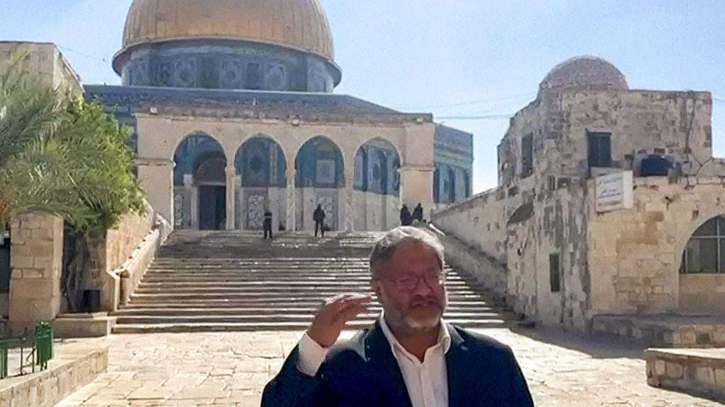 Itamar Ben-Gvir stands wearing a white dress shirt and black suit jacket, motioning with one hand and speaking, in front of the blue-tiled and gold-domed al-Aqsa mosque.