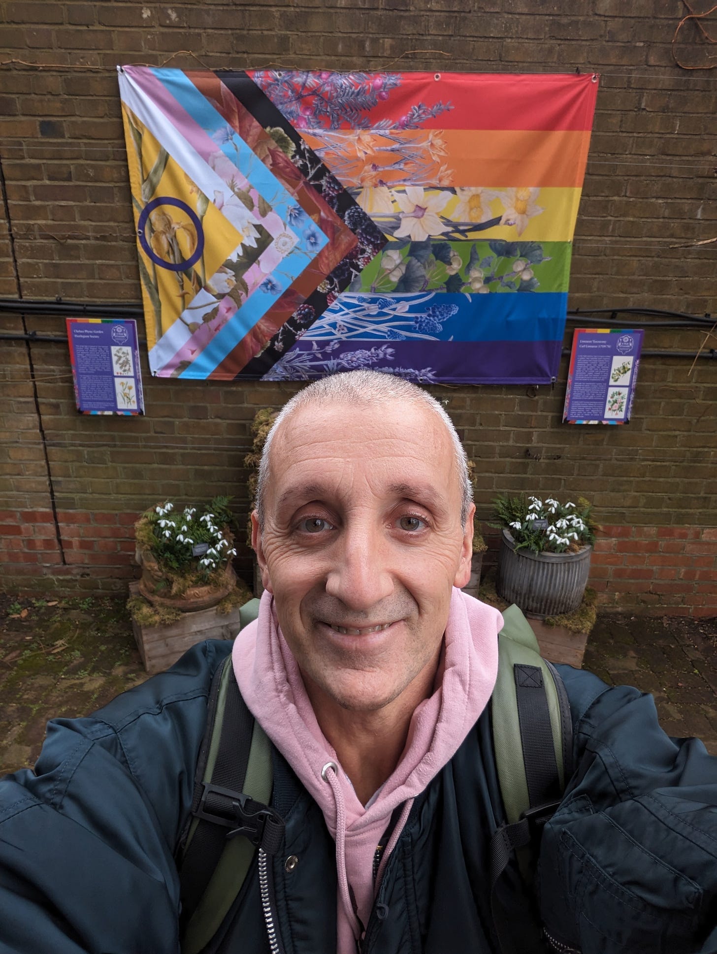 A man with short greying hair and kind eyes smiles at the camera. Behind him is a version of the progress flag with plants added to it, at Chelsea Physic Garden 2024 during LGBT History Month A man with short greying hair and kind eyes smiles at the camera. Behind him is a version of the progress flag with plants added to it, at Chelsea Physic Garden 2024 during LGBT History Month