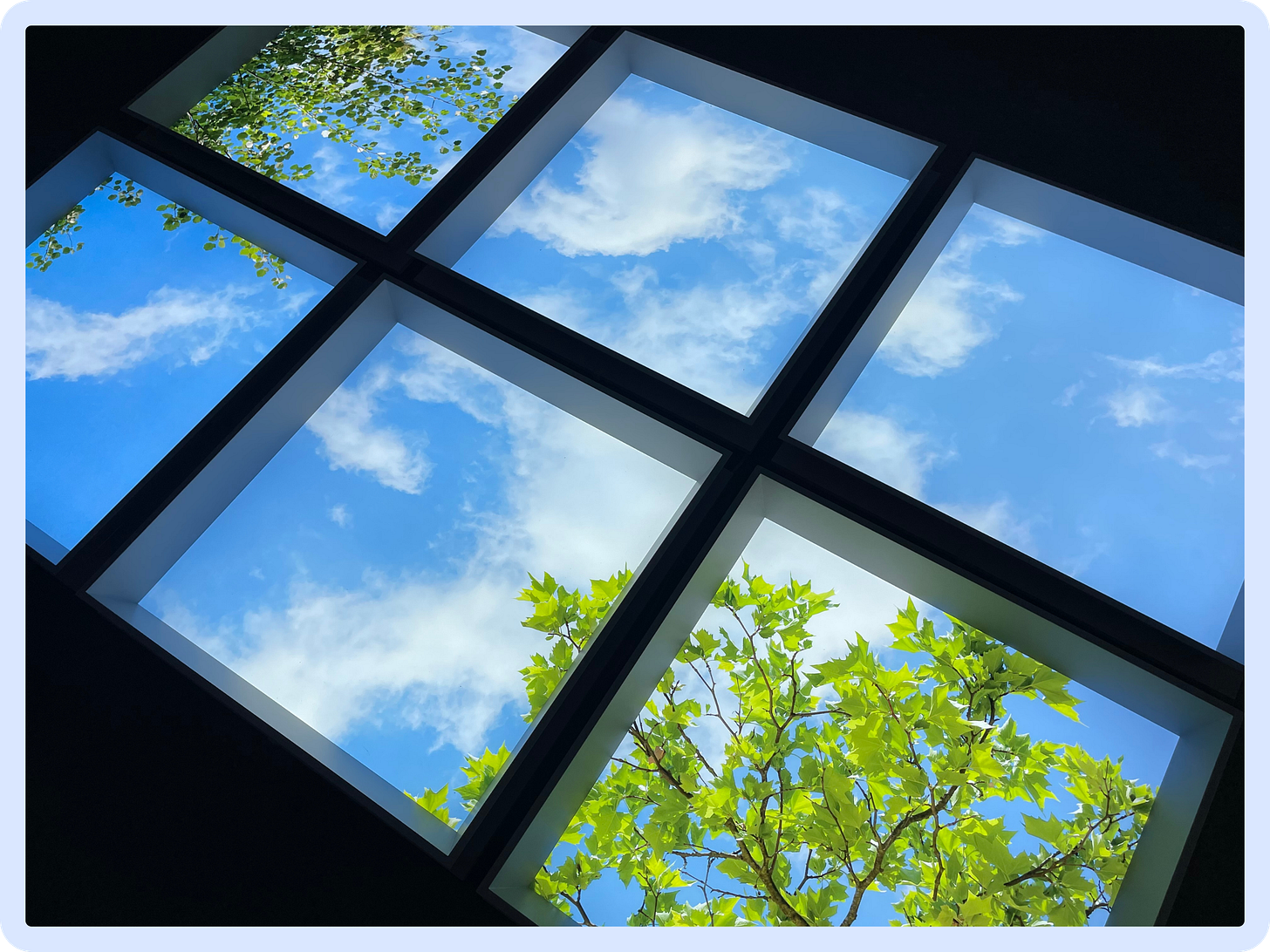 A photo of an installation of a faux-skylight laid flat on the floor, through which we can see a bright blue sky, wispy clouds, and luminous green trees. A photo of an installation of a faux-skylight laid flat on the floor, through which we can see a bright blue sky, wispy clouds, and luminous green trees.