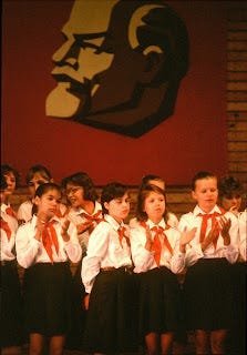 a class of kids in foreign school uniforms with a portrait of Stalin behind them