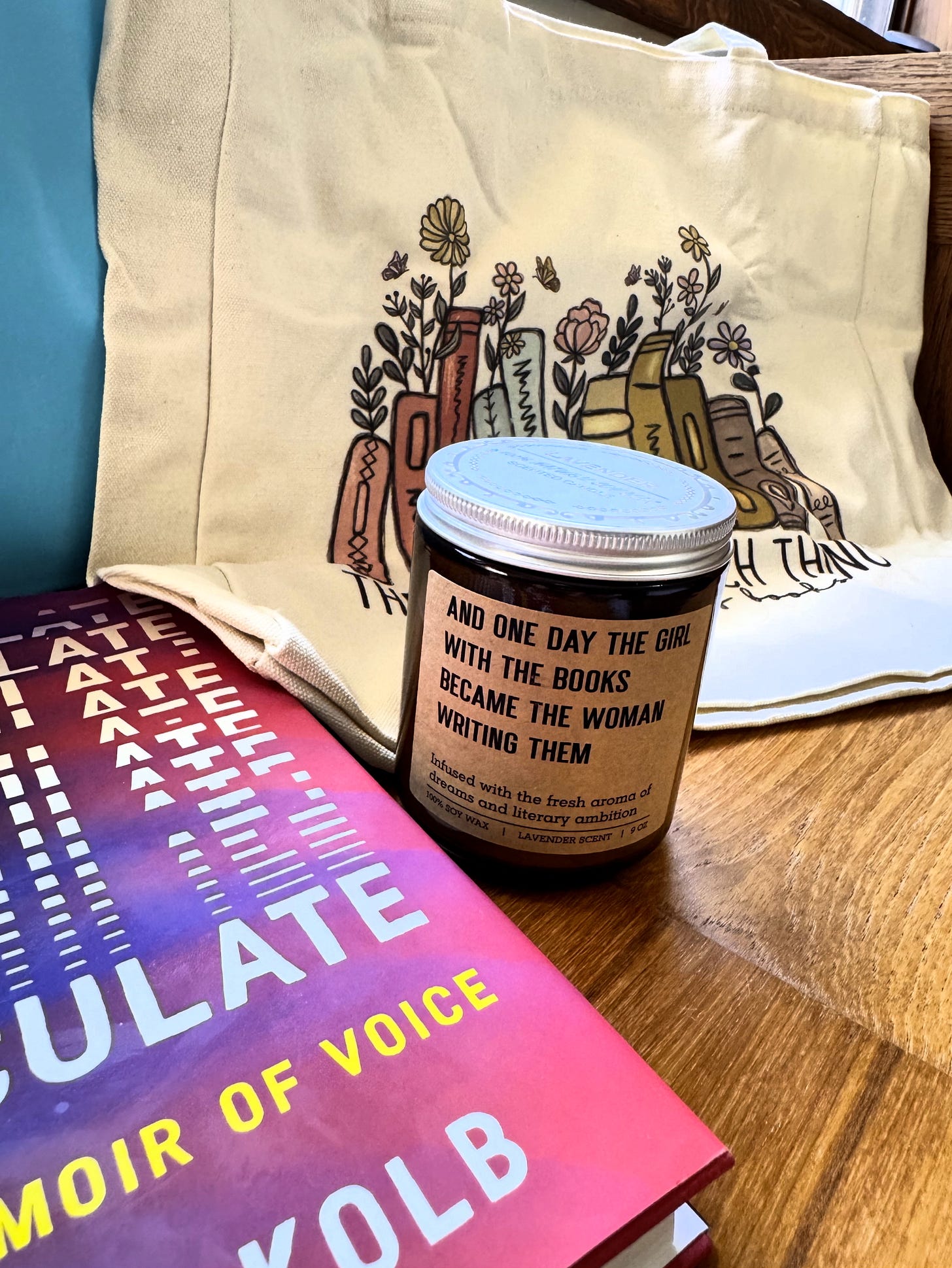 Photo featuring a lavender scented candle, with a side decal that reads: "And one day the girl with the books became the woman writing them." A hardcover of my book ARTICULATE is in the foreground.