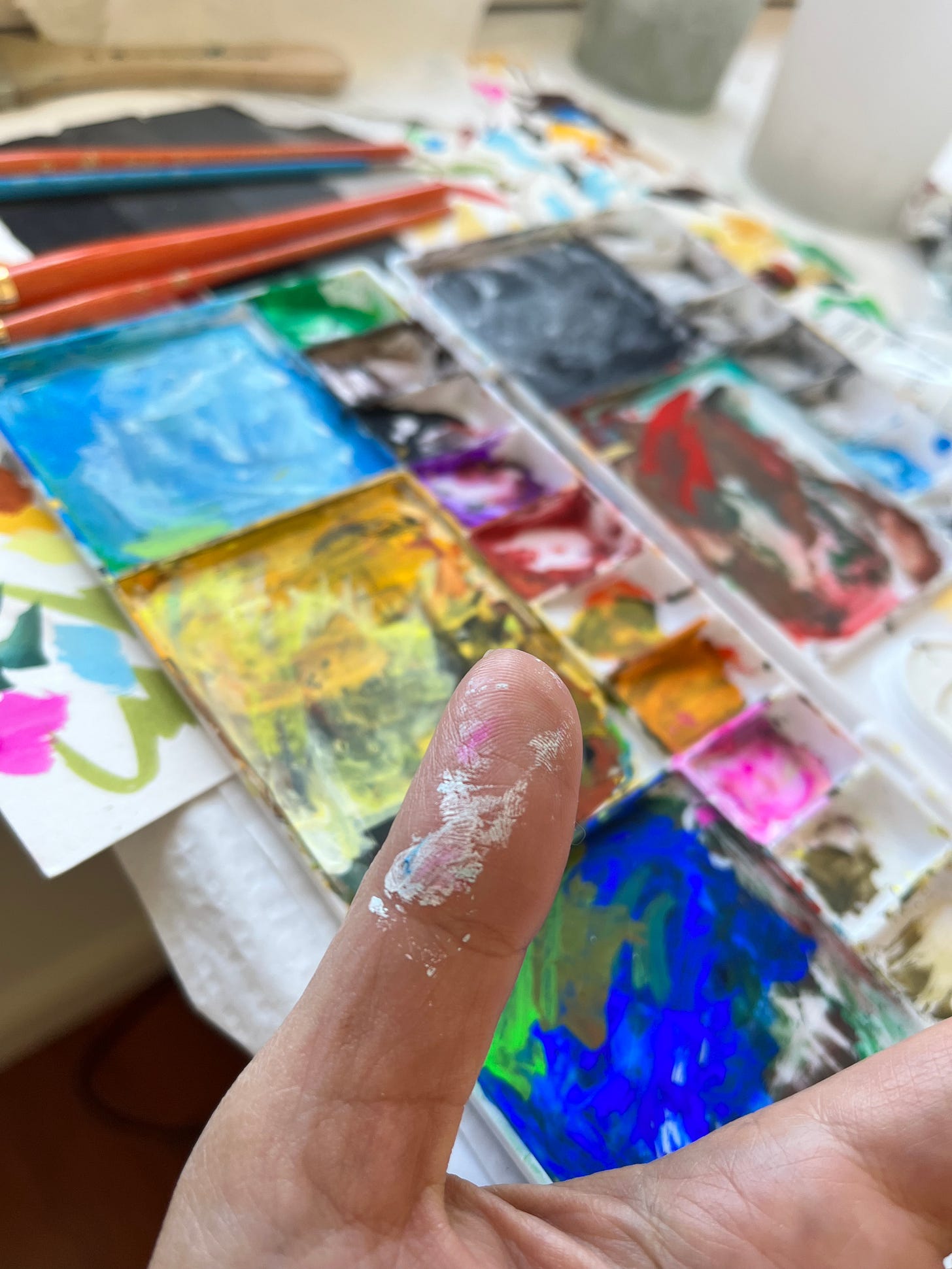 image: close-up of a painter's hand with fingers tainted with paint, in the background, colourful painting palette on the table.
