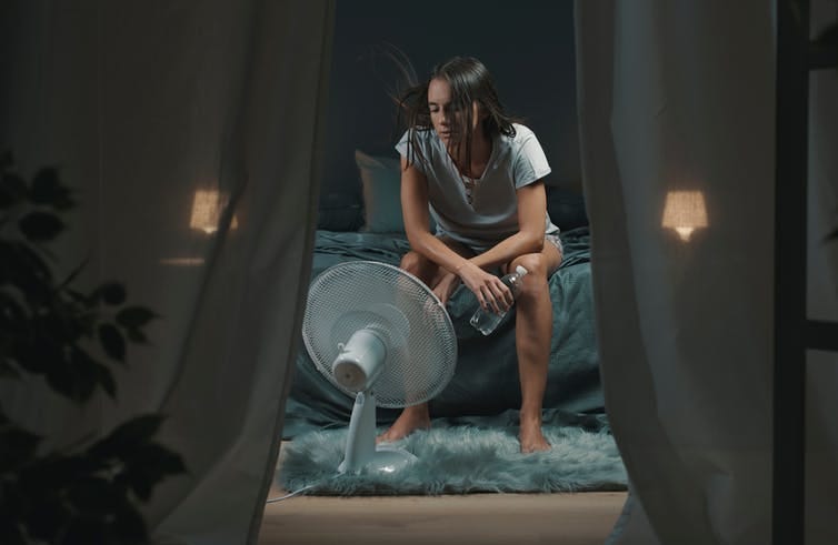 A woman sits on a bed in front of an electrical fan. A woman sits on a bed in front of an electrical fan.