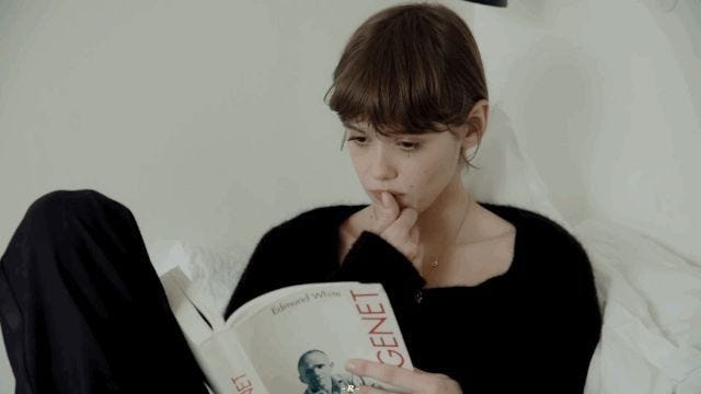 This may contain: a woman sitting on a bed reading a book with her hand under her chin and looking at the camera