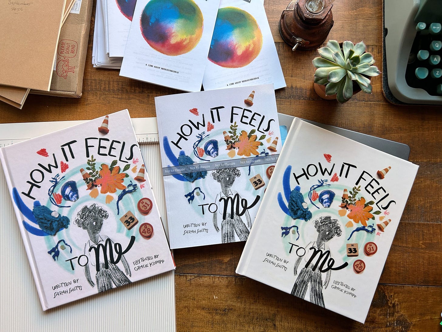 Three proofs of How it Feels to Me with a hand drawn figure surrounded by a halo of colorful objects. My typewriter, a succulent, and a stack of Spectrum zines are also visible.