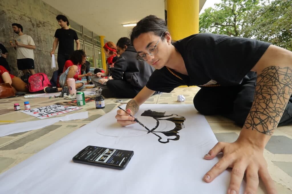 Student paints a protest sign on paper laid on the ground, with others nearby making banners and posters, one reading "Vote 13 Student paints a protest sign on paper laid on the ground, with others nearby making banners and posters, one reading "Vote 13
