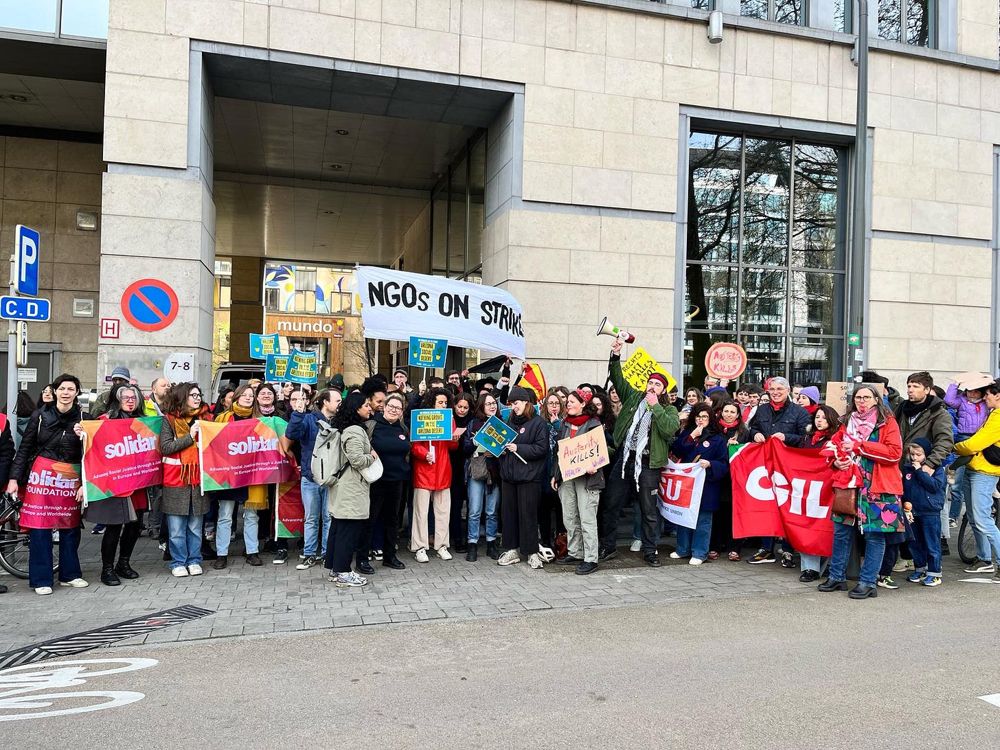 Group of people in front of an office building holding up signs that say 'Solidar' and 'austerity kills' and a big banner that says 'NGO workers on strike' in black and white. A man is talking into a megaphone