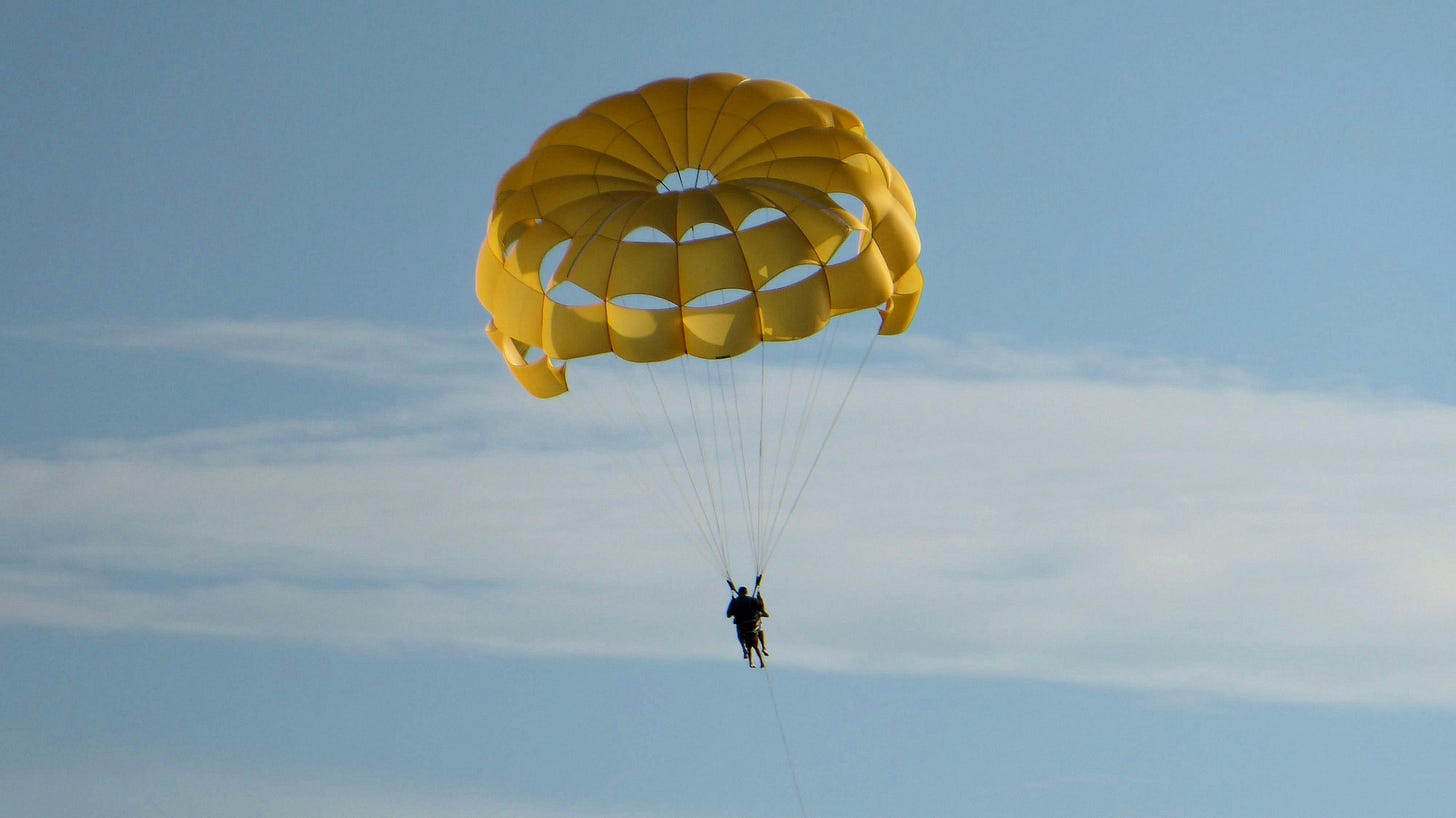 A yellow parachute, open against a blue sky with thin white clouds, carries a person safely to the ground.