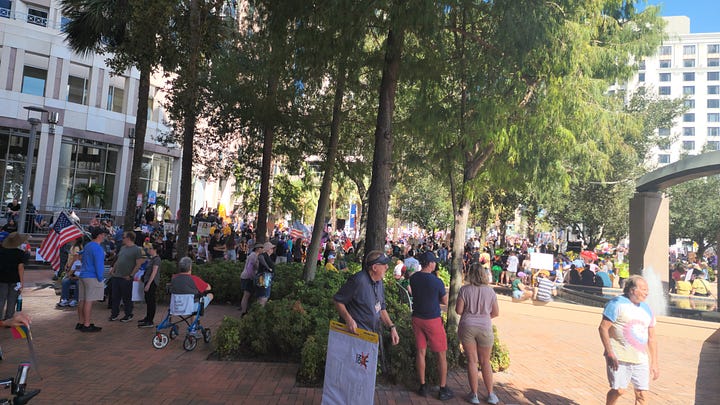 20251018_114518.jpg: A close-up of a hand-drawn protest sign reading "NO KINGS!!" and "NO WEENIES ALLOWED," featuring a crossed-out Burger King logo and a drawing of SpongeBob SquarePants.  20251018_113815.jpg: A wide shot of a large, sunny protest in a public plaza, showing crowds gathered around a modern arched sculpture and a water fountain.  20251018_113805.jpg: A crowd of protestors gathered on a brick plaza under a canopy of trees, with some people holding American flags and various signs.  20251018_110151 - Copy.jpg: A person in an inflatable blue pterodactyl costume stands at the protest, next to a person in a wheelchair holding a sign that reads, "DECLARE INDEPENDENT. FLUSH THE RED/BLUE WAR!"  20251018_113823.jpg: A wide view of the protest centered on a large, modern arched sculpture over a fountain, with a large crowd and many signs visible in the background.