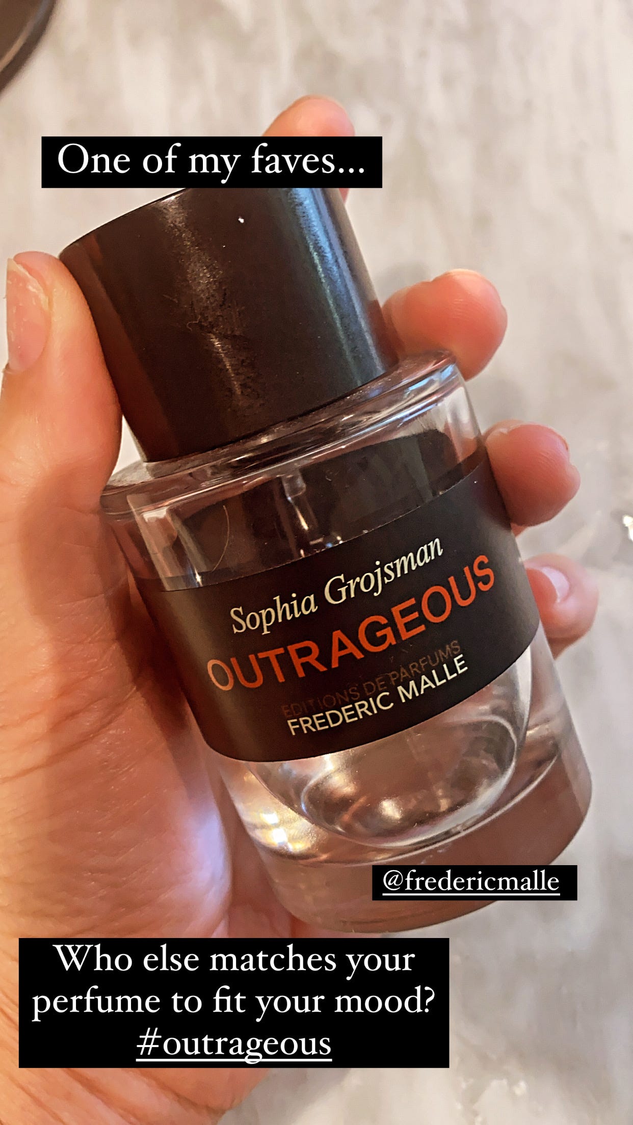 A hand holding a bottle of Editions de Parfums Frederic Malle "Outrageous" perfume against a marble surface.