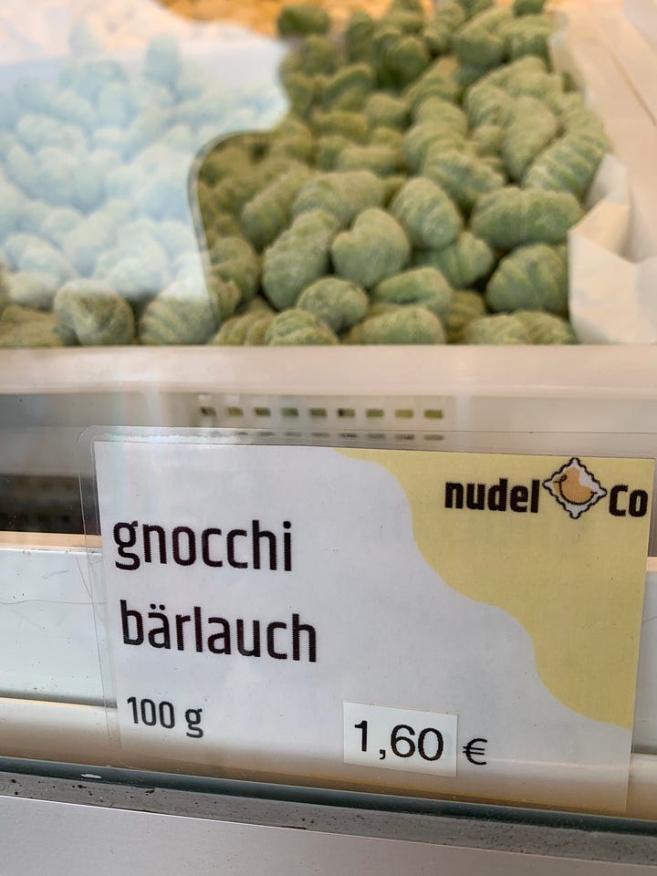 top left to bottom right: image of two spring raspberry colored cocktails with olives and a thick tubular pasta "straw", containers of white asparagus for sale at 12.99Euro per kilo, barlauch gnocchi (green)with price, cheerful red Karl'sStrawberries kiosk