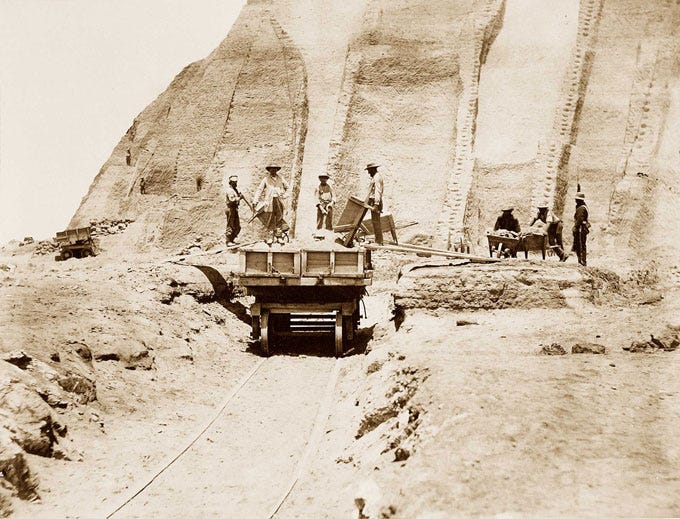 This image from 1865 shows workers excavating a mountain of bird poop. Some are pushing wheelbarrows full and others are emptying wheelbarrows into a wagon that is on tracks leading away from the dung heap.