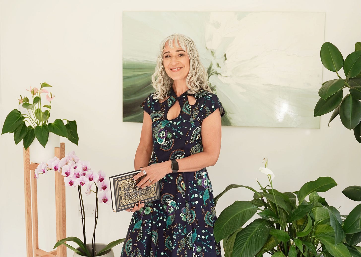 Sanjida in a blue dress holding a book, standing in her office, surrounded by plants and with a white and green abstract painting behind her.