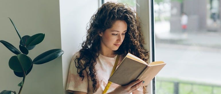 a woman sitting on a window sill reading a book next to two dogs a woman sitting on a window sill reading a book next to two dogs