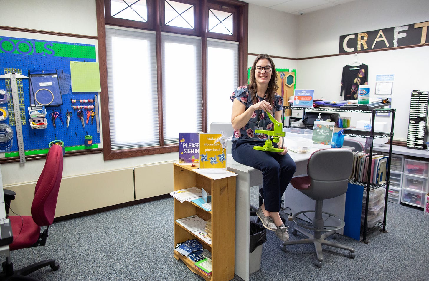 A librarian sits on a tabletop surrounded by craft and maker project supplies, lit by a large window in a library makerspace. A librarian sits on a tabletop surrounded by craft and maker project supplies, lit by a large window in a library makerspace.