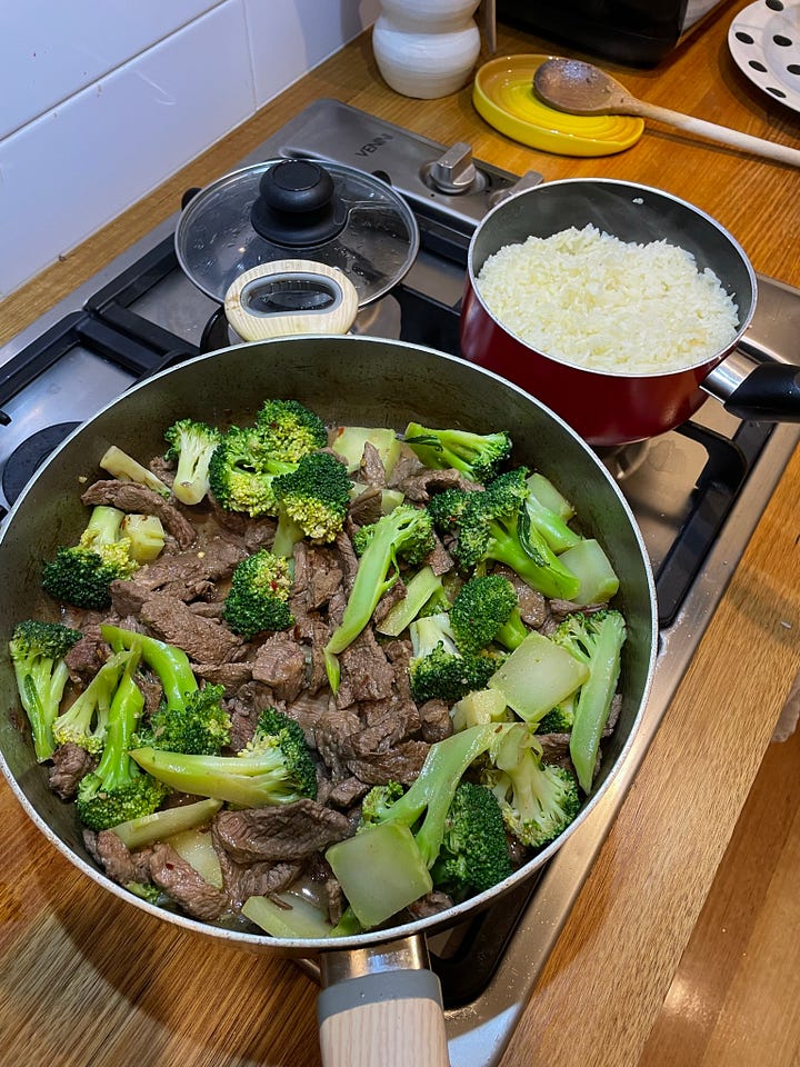 Saucepan full of beef and broccoli, garlic rice in a smaller pot alongside. A table lit with a lamp showing two slices of chocolate cake with salted chocolate icing.