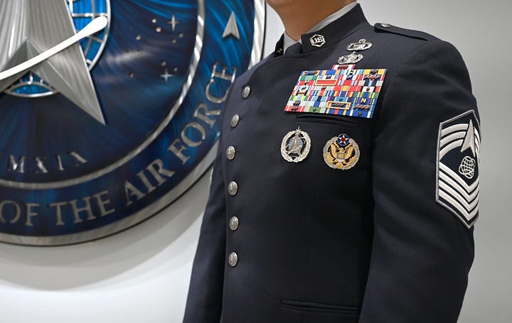 A smiling U.S. Space Force service member walks down a bright, polished hallway lined with wood paneling and framed portraits of military leaders. She wears a dark-blue dress uniform with silver buttons, senior enlisted chevrons, a chest full of ribbons, and a Space Force insignia, paired with a gray skirt. Fluorescent ceiling lights and glossy tile floors lead toward glass doors at the end of the corridor.