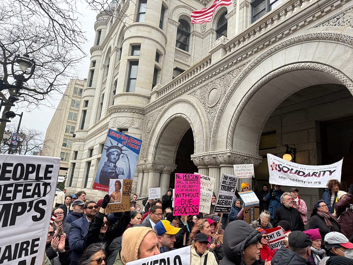 A crowd in front of a federal building with an arched entryway. THey are holding signs, including a State of LIberty made up as Rosie the Riveter saying "STOP THE ABUSE OF POWER" and quoting the Due Process clause of the Constitution the picture on the left and "Resist Fascism, Dump Trump," an upside-down US flag and "Drop The Charges" on the right.