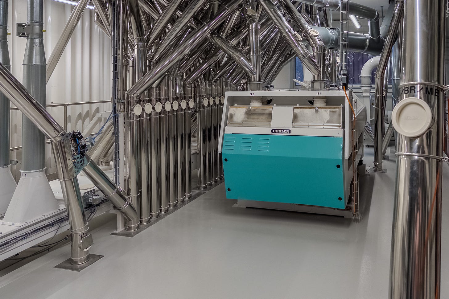 Blick in einen technischen Prozessraum einer Industrie-Mühle: Zahlreiche senkrechte und schräge Edelstahlrohre verlaufen durch den Raum und führen zu einer türkis-weissen Bühler-Maschine. Wahrscheinlich zeigt das Bild die Sichterei oder einen Verteilbereich, in dem das Mahlgut nach Fraktionen getrennt und über Luftförderung weitertransportiert wird. Blick in einen technischen Prozessraum einer Industrie-Mühle: Zahlreiche senkrechte und schräge Edelstahlrohre verlaufen durch den Raum und führen zu einer türkis-weissen Bühler-Maschine. Wahrscheinlich zeigt das Bild die Sichterei oder einen Verteilbereich, in dem das Mahlgut nach Fraktionen getrennt und über Luftförderung weitertransportiert wird.
