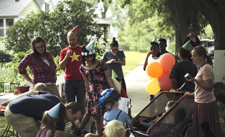 People in party hats gather on a sidewalk, getting ready to take a walking tour of their neighborhood.