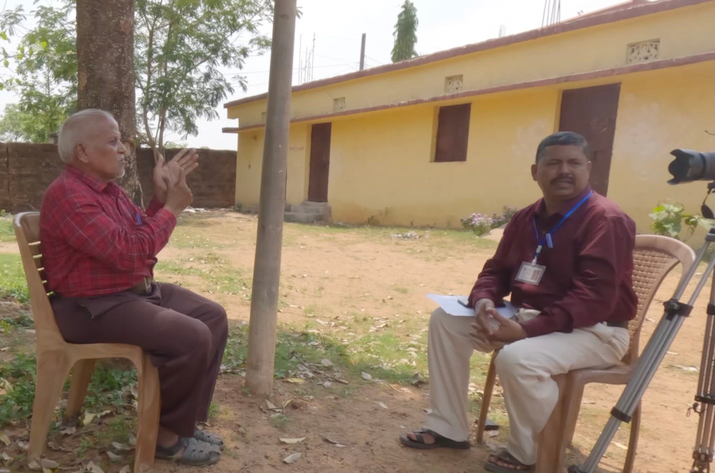 A MAN WITH GRAY HAIR MAKES SIGN LANGUAGE GESTURES TO ANOTHER. A CAMERA POINTS TOWARD HIM.