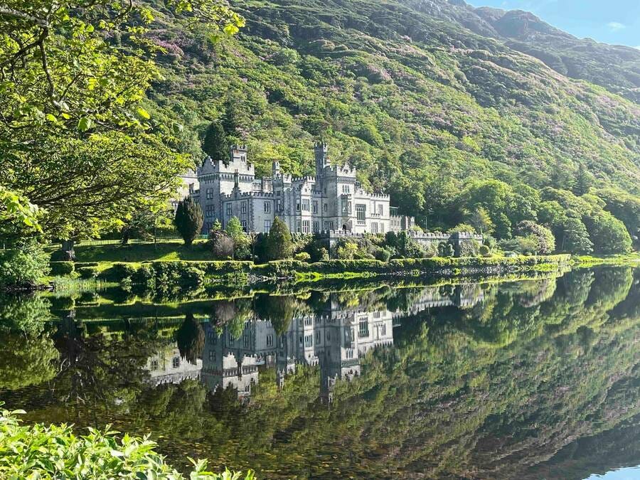 A white abbey on a green lawn, with castle-like features, sitting at the base of a hill covered in trees, with a lake reflecting the scene in its waters.