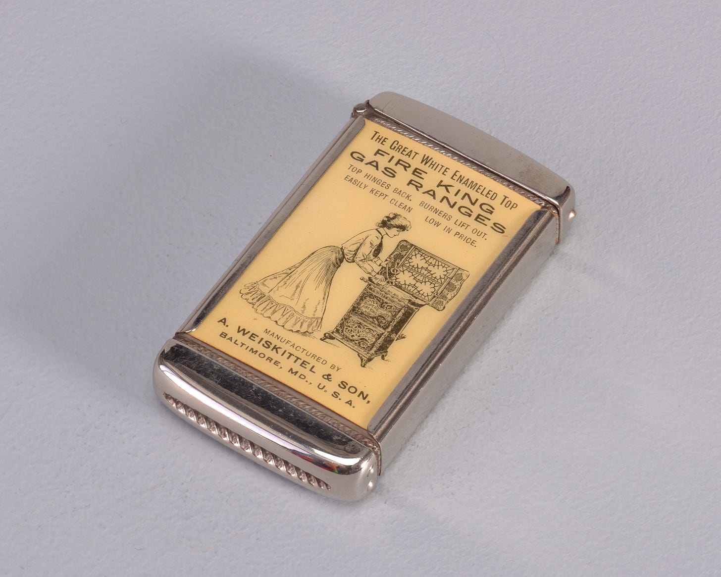 Rectangular, curved top and bottom, featuring black printed decoration of female in blouse, tie, long skirt, and upswept hair cleaning beneath range top, inscribed above "The Great White Enameled Top. Fire King Gas Ranges. Top Hinges Back. Burners Lift Out. Easily Kept Clean. Low in Price." Inscribed below "Manufactured By A. Weiskittel & Son, Baltimore, MD., U.S.A." Reverse features hot plate, inscribed above "A Match Is All The Kindling Required Where Fire King Gas Stoves And Ranges Are Used." Inscribed below "New Fire King Hot Plate With Hinged Top Gratings And Removable Drilled Burners." Inscribed in fine print below "The Whitehead & Hoag Co., Newark, N.J." Lid hinged on side. Striker on bottom.