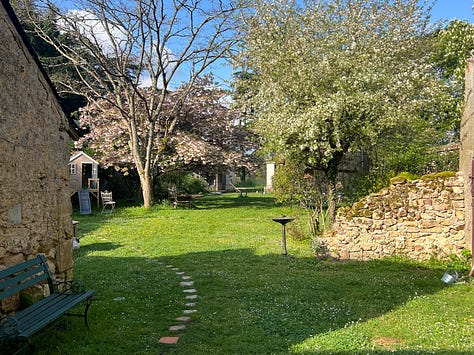A collage of the grounds around the rented farmhouse, including an iron gate set into an ivy-covered stone wall, a garden path, a courtyard strung with bunting at dusk, two small children watching a rainbow from a doorway, pink cyclamen carpeting the ground beneath a large tree, and children walking down tree-lined country paths.