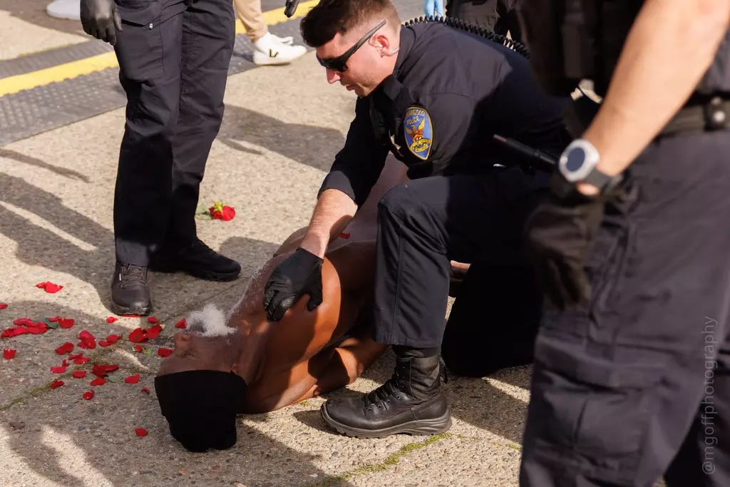 Police officers restrain a nude Black man lying on the pavement, surrounded by scattered red flower petals, during a protest at the Bob Weir memorial in San Francisco. One officer is kneeling beside him while others stand nearby. Police officers restrain a nude Black man lying on the pavement, surrounded by scattered red flower petals, during a protest at the Bob Weir memorial in San Francisco. One officer is kneeling beside him while others stand nearby.