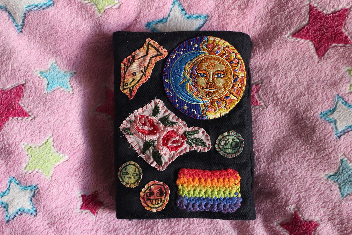 the back of a black, clothbound sketchbook with several colorful patches sewn to it, including roses, a fish, a rainbow, faces, and a sun and moon