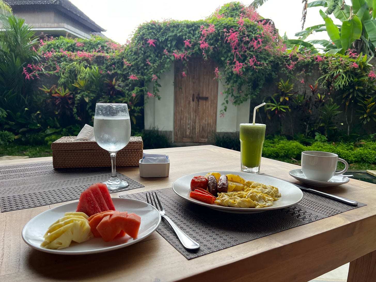A breakfast tray with eggs, potatoes, a blistered tomato, cut fruit fanned out, a green juice and coffee.