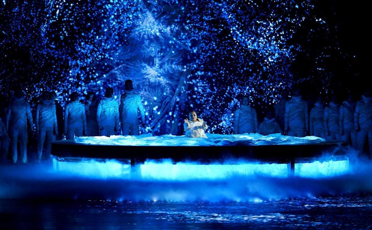 A disabled femme with osteogenesis imperfecta sits alone on a floating stage surrounded by performers in the shadowy background. Light projections cast brilliant patterns that look like particles of ice or snow. A disabled white femme with osteogenesis imperfecta sits alone on a floating stage surrounded by performers in the shadowy background. Light projections cast brilliant patterns that look like particles of ice or snow.