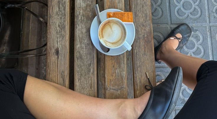 This may contain: two people sitting at a wooden table with cups of coffee on it and one person's legs crossed