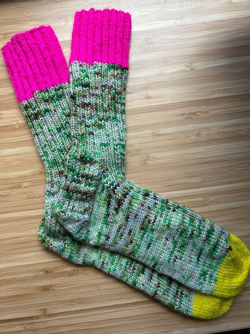 A pair of handknit socks with bright pink ribbing on the top and a marled green yarn for the main color.